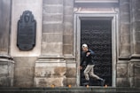 A person wearing rollerblades performs a maneuver on a stone surface in front of an ornately decorated black door set in a stone wall. There are small orange traffic cones arranged on the ground. The wall features a mounted plaque with inscriptions and an embossed figure.