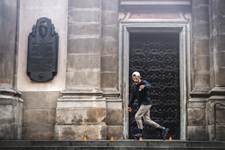 A person wearing rollerblades performs a maneuver on a stone surface in front of an ornately decorated black door set in a stone wall. There are small orange traffic cones arranged on the ground. The wall features a mounted plaque with inscriptions and an embossed figure.