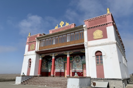 A traditional Asian-style building with ornate decorations, featuring vibrant red, gold, and white colors. The structure has detailed artwork and symbols on its facade, with a staircase leading up to the entrance. The clear blue sky forms the backdrop, adding to the serene and tranquil atmosphere.