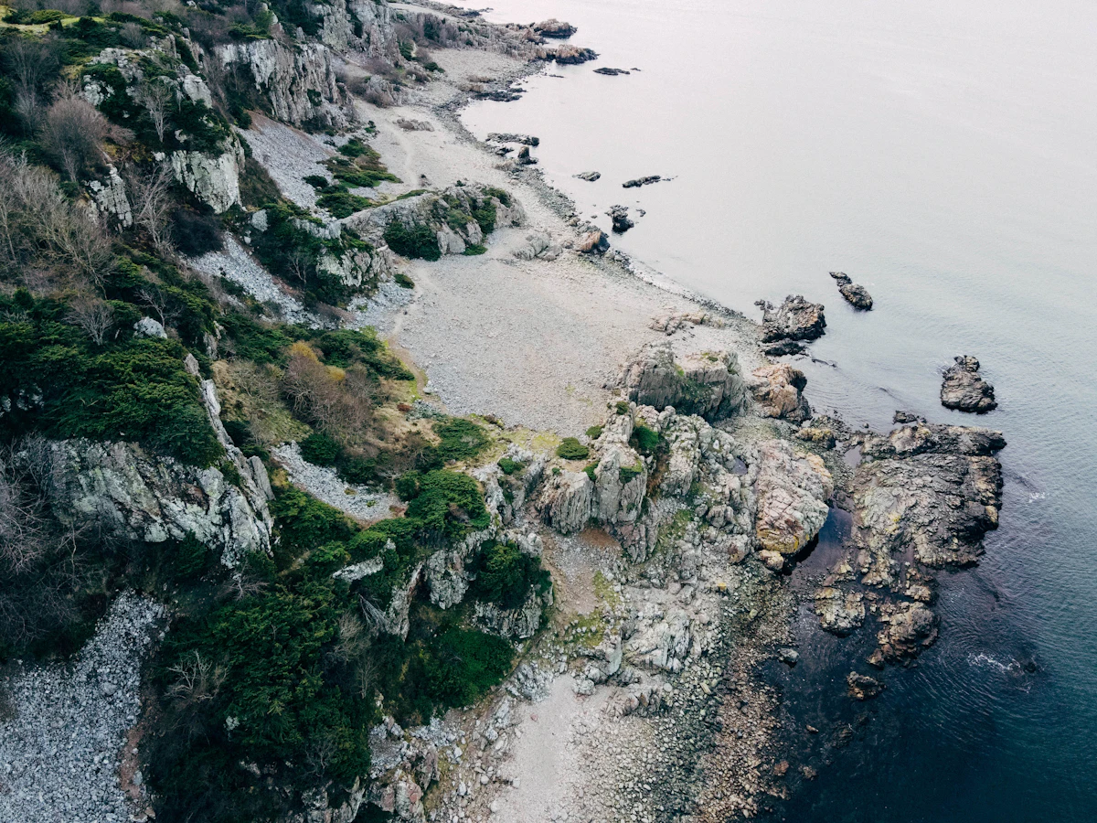 An aerial view of a steep, scrubby headland and a pebble beach on the coast near Båstad
