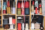 Multiple wooden shelves filled with neatly organized spools of colorful ribbons and cords of varying textures and thicknesses. The assortment has a mix of solid colors and patterns, providing a vibrant and eclectic appearance. Labels can be seen on some shelves, possibly indicating different categories or types of materials.