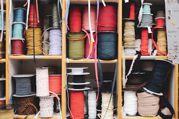 Multiple wooden shelves filled with neatly organized spools of colorful ribbons and cords of varying textures and thicknesses. The assortment has a mix of solid colors and patterns, providing a vibrant and eclectic appearance. Labels can be seen on some shelves, possibly indicating different categories or types of materials.