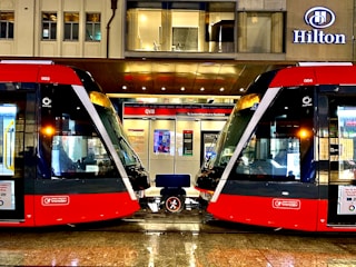 Two modern red trams are positioned side by side on a wet platform in an urban setting. Above the platform is a building with a sign displaying the name Hilton. The platform is well-lit and details of the tram interiors can be seen through the large windows. A bench is situated between the trams.