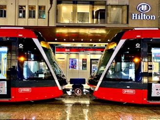 Two modern red trams are positioned side by side on a wet platform in an urban setting. Above the platform is a building with a sign displaying the name Hilton. The platform is well-lit and details of the tram interiors can be seen through the large windows. A bench is situated between the trams.