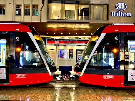 Two modern red trams are positioned side by side on a wet platform in an urban setting. Above the platform is a building with a sign displaying the name Hilton. The platform is well-lit and details of the tram interiors can be seen through the large windows. A bench is situated between the trams.