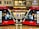 Two modern red trams are positioned side by side on a wet platform in an urban setting. Above the platform is a building with a sign displaying the name Hilton. The platform is well-lit and details of the tram interiors can be seen through the large windows. A bench is situated between the trams.