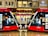 Two modern red trams are positioned side by side on a wet platform in an urban setting. Above the platform is a building with a sign displaying the name Hilton. The platform is well-lit and details of the tram interiors can be seen through the large windows. A bench is situated between the trams.