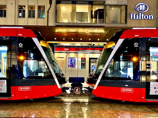 Two modern red trams are positioned side by side on a wet platform in an urban setting. Above the platform is a building with a sign displaying the name Hilton. The platform is well-lit and details of the tram interiors can be seen through the large windows. A bench is situated between the trams.