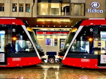 Two modern red trams are positioned side by side on a wet platform in an urban setting. Above the platform is a building with a sign displaying the name Hilton. The platform is well-lit and details of the tram interiors can be seen through the large windows. A bench is situated between the trams.