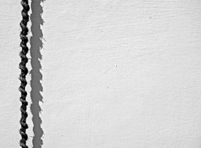 A black rope casts a shadow on a light textured surface, creating a striking contrast between the dark rope and the bright background. The shadow follows the irregular contours of the rope, adding a sense of depth and dynamic curvature.