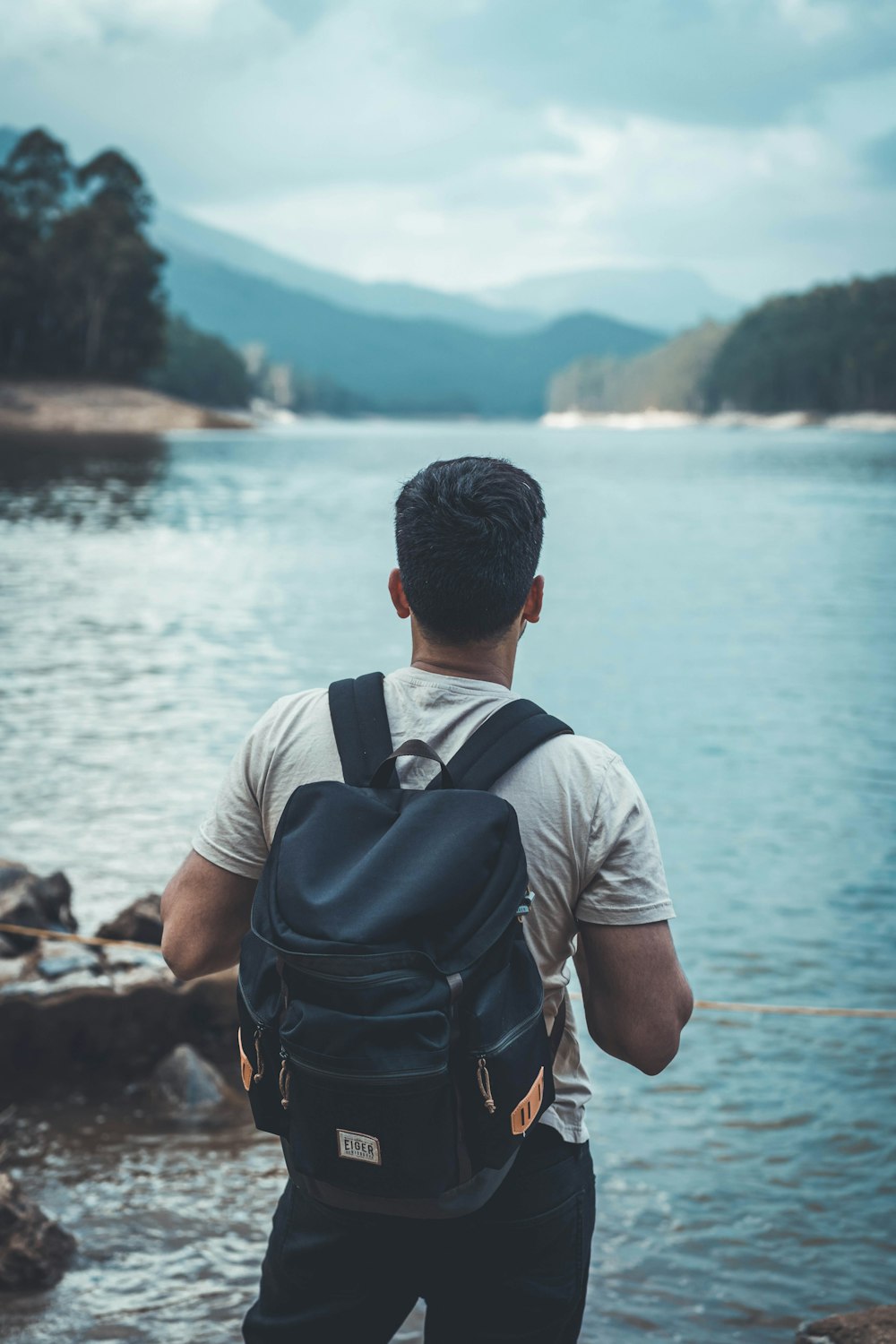 Homme en chemise grise et sac à dos noir debout sur le rivage rocheux pendant la journée