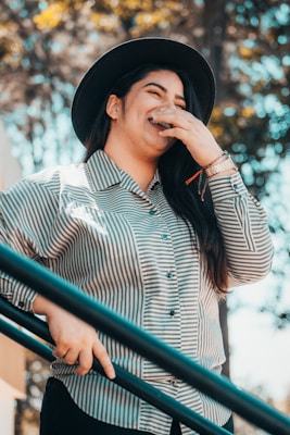 A person wearing a black hat and striped shirt is laughing while standing near a railing, with their hand covering their mouth. Sunlight filters through the trees in the background, creating a warm and relaxed atmosphere.