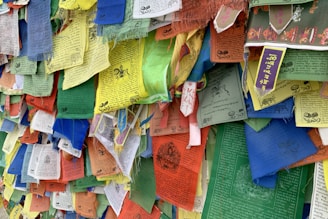 A colorful assortment of rectangular prayer flags with various inscriptions and images, featuring multiple colors such as blue, yellow, red, green, and white. The flags are frayed at the edges, indicating exposure to wind and weather. They are closely packed together, creating a vibrant tapestry effect.
