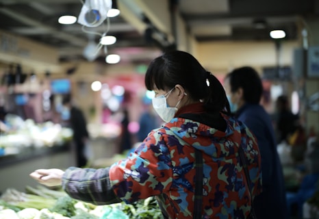 A person wearing a colorful, patterned jacket and a facemask is shopping at a market. The setting is indoors, with fluorescent lighting. The person is reaching out towards a selection of fresh vegetables, including leafy greens and cabbages. Other shoppers and stalls are visible in the background, slightly out of focus.