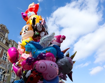 A vibrant cluster of colorful balloons featuring various cartoon characters, including animals and fantastical creatures, set against a backdrop of a clear blue sky with some clouds. The balloons are arranged in a towering display rising above the urban street scene.