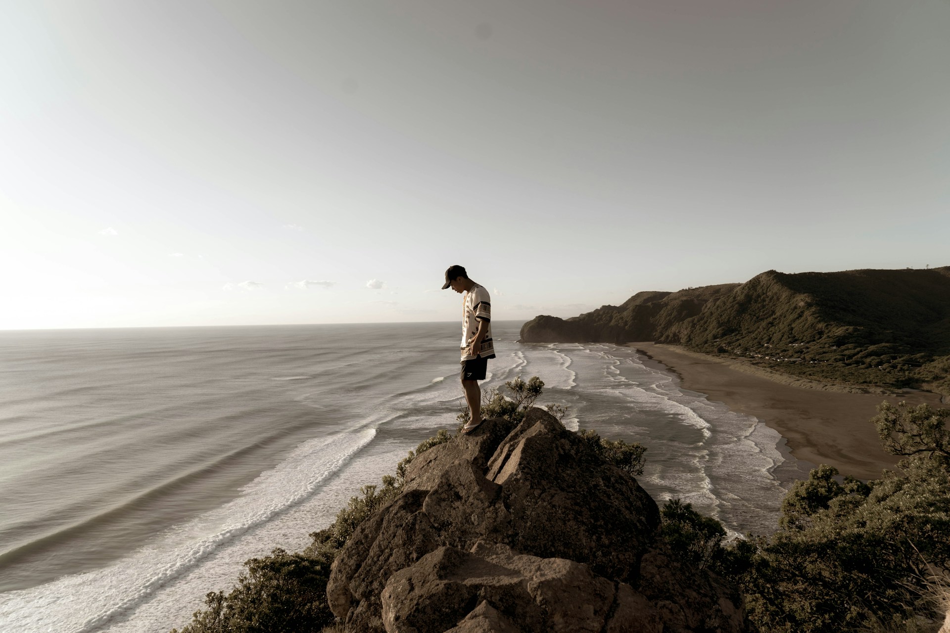 man in white t-shirt and black shorts standing on rock formation near sea during daytime
