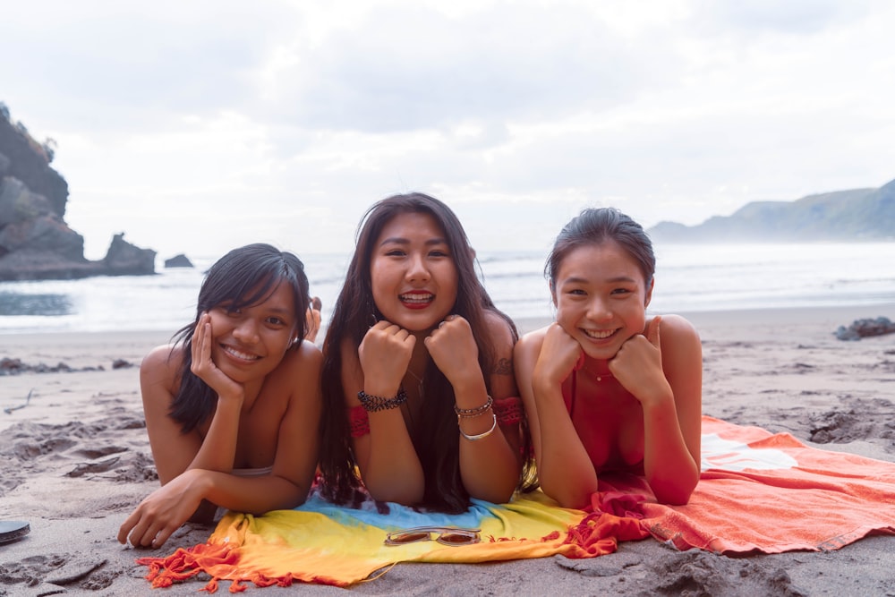 3 women in bikini sitting on red textile on beach during daytime