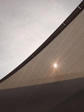 A close-up view of a fabric canopy, with sunlight creating a bright spot on the textured surface. The canopy is stretched taut, with visible seams or supports. The sky in the background is overcast, adding a muted tone to the overall scene.