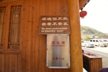 A wooden building with a window on the left and a metal fire hydrant box mounted on the wall. Above the fire hydrant box, there is a wooden sign with both Chinese and English text, stating 'No clean environment, no beautiful view!' In the background, there are parked cars and a glimpse of a hilly landscape.