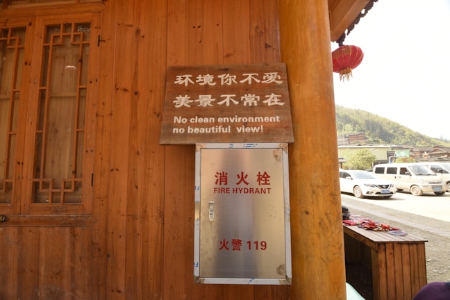 A wooden building with a window on the left and a metal fire hydrant box mounted on the wall. Above the fire hydrant box, there is a wooden sign with both Chinese and English text, stating 'No clean environment, no beautiful view!' In the background, there are parked cars and a glimpse of a hilly landscape.