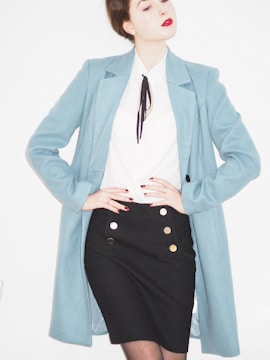 A person is wearing a stylish outfit featuring a long light blue coat over a white blouse with a black tie or accessory at the collar. The outfit is paired with a fitted black skirt adorned with gold buttons. The individual has red lipstick and their hair is styled in an updo.