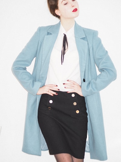 A person is wearing a stylish outfit featuring a long light blue coat over a white blouse with a black tie or accessory at the collar. The outfit is paired with a fitted black skirt adorned with gold buttons. The individual has red lipstick and their hair is styled in an updo.