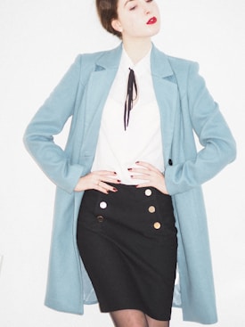 A person is wearing a stylish outfit featuring a long light blue coat over a white blouse with a black tie or accessory at the collar. The outfit is paired with a fitted black skirt adorned with gold buttons. The individual has red lipstick and their hair is styled in an updo.