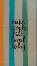 A motivational message is painted on a brick wall, featuring hand-lettered text that reads 'make people feel loved today.' The background consists of vertical stripes in various colors.