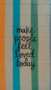 A motivational message is painted on a brick wall, featuring hand-lettered text that reads 'make people feel loved today.' The background consists of vertical stripes in various colors.