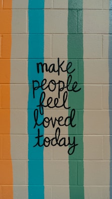 A motivational message is painted on a brick wall, featuring hand-lettered text that reads 'make people feel loved today.' The background consists of vertical stripes in various colors.