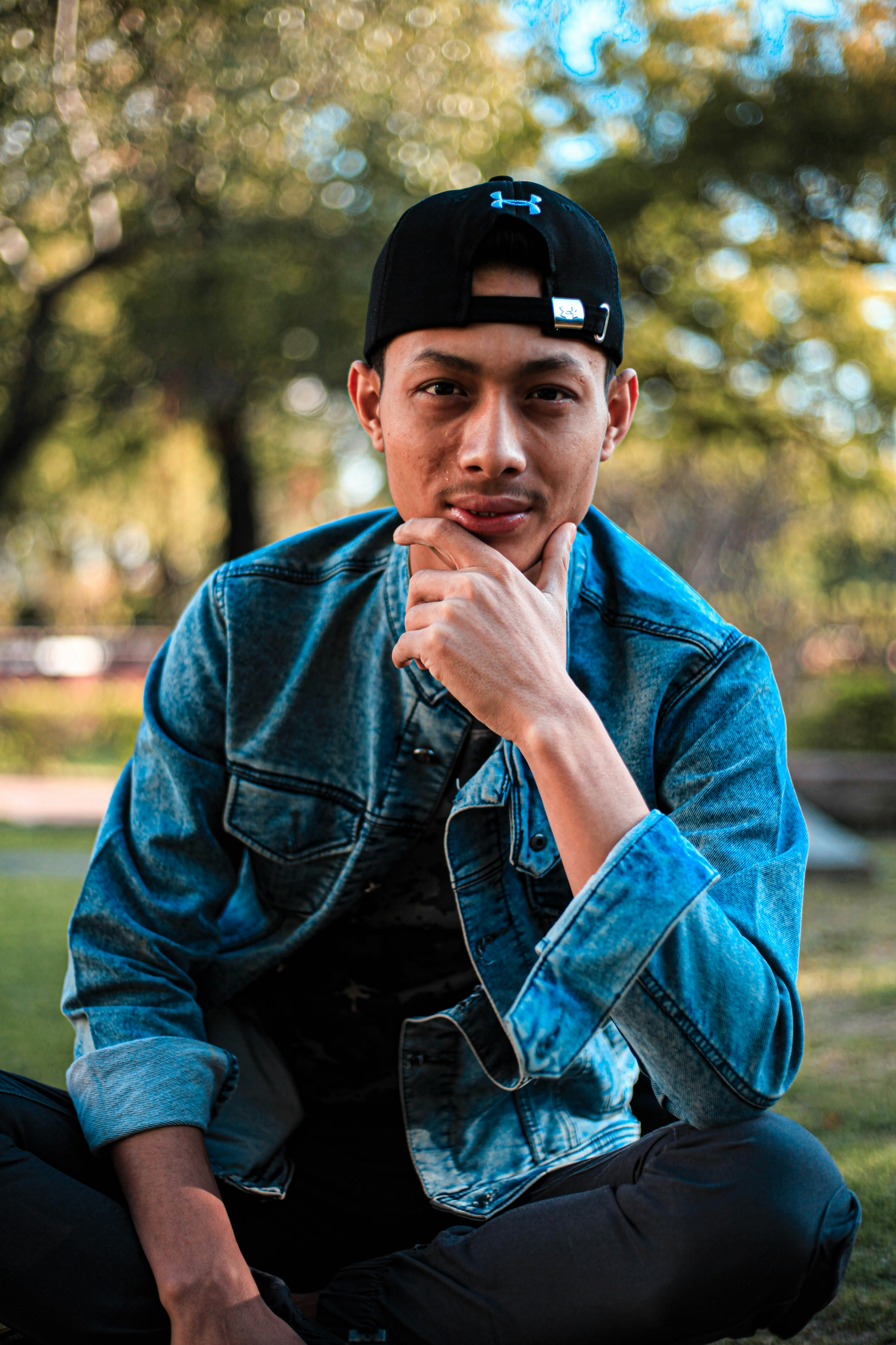 昼間に緑の草原に座っている青いデニム ジャケットと黒のフィット キャップの男の写真 Unsplashで見つける衣類の無料写真