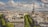 A panoramic view of a cityscape featuring the Eiffel Tower prominently in the center, surrounded by historical buildings with classic Haussmann-style architecture. The scene includes tree-lined boulevards and a partly cloudy sky above the urban landscape.