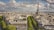 A panoramic view of a cityscape featuring the Eiffel Tower prominently in the center, surrounded by historical buildings with classic Haussmann-style architecture. The scene includes tree-lined boulevards and a partly cloudy sky above the urban landscape.