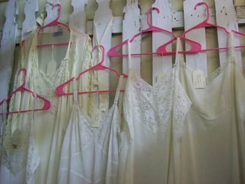 Several lacey white dresses or nightgowns are hanging on bright pink hangers against a white wooden fence. Each garment has a tag attached to it.
