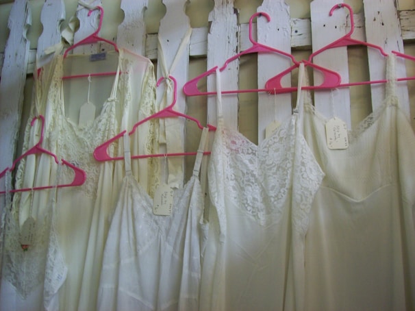 Several lacey white dresses or nightgowns are hanging on bright pink hangers against a white wooden fence. Each garment has a tag attached to it.