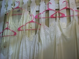 Several lacey white dresses or nightgowns are hanging on bright pink hangers against a white wooden fence. Each garment has a tag attached to it.