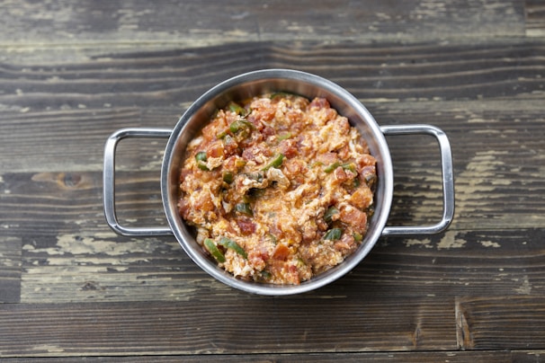 A stainless steel bowl filled with a mixture of scrambled eggs, tomatoes, and green peppers, placed on a rustic wooden surface.