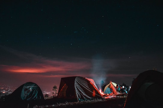 A nighttime camping scene with tents set up on a grassy area. The sky is clear with visible stars and a subtle glow from the horizon, possibly from city lights. A campfire is burning in the foreground, casting light on the tents and creating a warm, inviting atmosphere against the dark night.