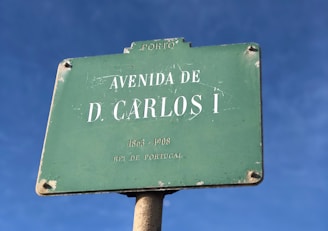 A green street sign with white text displaying 'Avenida de D. Carlos I'. The sign shows signs of wear with chipped paint and rust. The background is a clear blue sky.