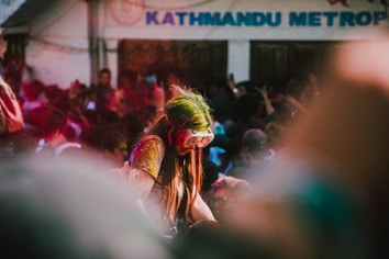 A person covered in colored powders stands amidst a large crowd. The individual wears goggles and their hair is streaked with bright green. The background is filled with people, all appearing to be enjoying a festive event, with the word 'KATHMANDU' partially visible on a building.