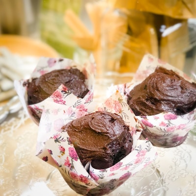 Three chocolate muffins are displayed on a decorative surface, each wrapped in paper with a floral design. The background is softly blurred, creating a warm and inviting atmosphere.