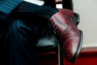 A close-up view of a polished brown leather brogue shoe worn by a person crossing their legs. The individual is dressed in a dark pinstripe suit, with both the shoe and suit conveying a formal and sophisticated style. The shoe features intricate perforated details typical of brogue designs.