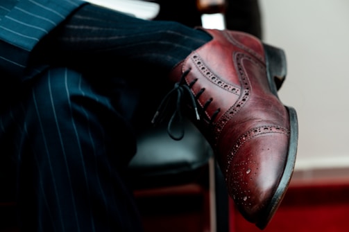 A close-up view of a polished brown leather brogue shoe worn by a person crossing their legs. The individual is dressed in a dark pinstripe suit, with both the shoe and suit conveying a formal and sophisticated style. The shoe features intricate perforated details typical of brogue designs.