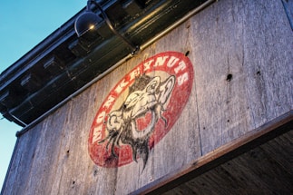A rustic wooden sign features an illustration of a monkey surrounded by a red oval background, with the text 'MONKEYNUTS' written in a bold, curving font. The sign is mounted under a black metal lamp and is affixed to a weathered wooden surface.