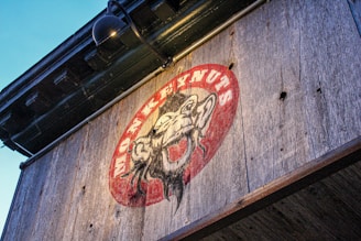 A rustic wooden sign features an illustration of a monkey surrounded by a red oval background, with the text 'MONKEYNUTS' written in a bold, curving font. The sign is mounted under a black metal lamp and is affixed to a weathered wooden surface.