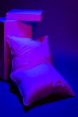 A pillow is positioned against geometric blocks under bold purple and blue lighting. The scene is minimalistic with a focus on shapes and colors.