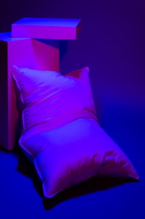 A pillow is positioned against geometric blocks under bold purple and blue lighting. The scene is minimalistic with a focus on shapes and colors.