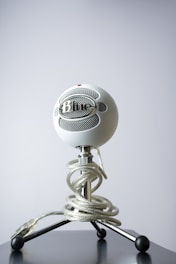 A white spherical microphone sits on a black, three-legged stand. The microphone is secured with a clear coiled cable wrapped around the stand. The background is a plain gray, providing contrast to the microphone's intricate design.