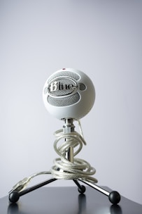 A white spherical microphone sits on a black, three-legged stand. The microphone is secured with a clear coiled cable wrapped around the stand. The background is a plain gray, providing contrast to the microphone's intricate design.
