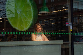 A large green lime slice graphic is displayed on the glass window of a shop. Inside, a person with shoulder-length hair and an orange striped shirt leans slightly forward, appearing cheerful. The interior features shelves and items typical of a store setting, and the lighting creates reflections on the glass.